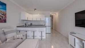 A modern kitchen with white cabinets, a marble countertop, and a built-in oven. There is a dining area with a white table and bar stools. A flat-screen TV is mounted on the wall. - Kaora Ocean View Apartment (Costa Adeje)