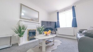 A modern living room with a white sofa, a white coffee table with fruit, a fireplace, a television, and a large window with curtains. - Sleeps 5 - Private Garden- Free Parking (Corby)
