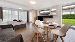 A modern hotel room with a bed, a dining area with a table and chairs, a kitchen area, and large windows offering a view of the outdoors.