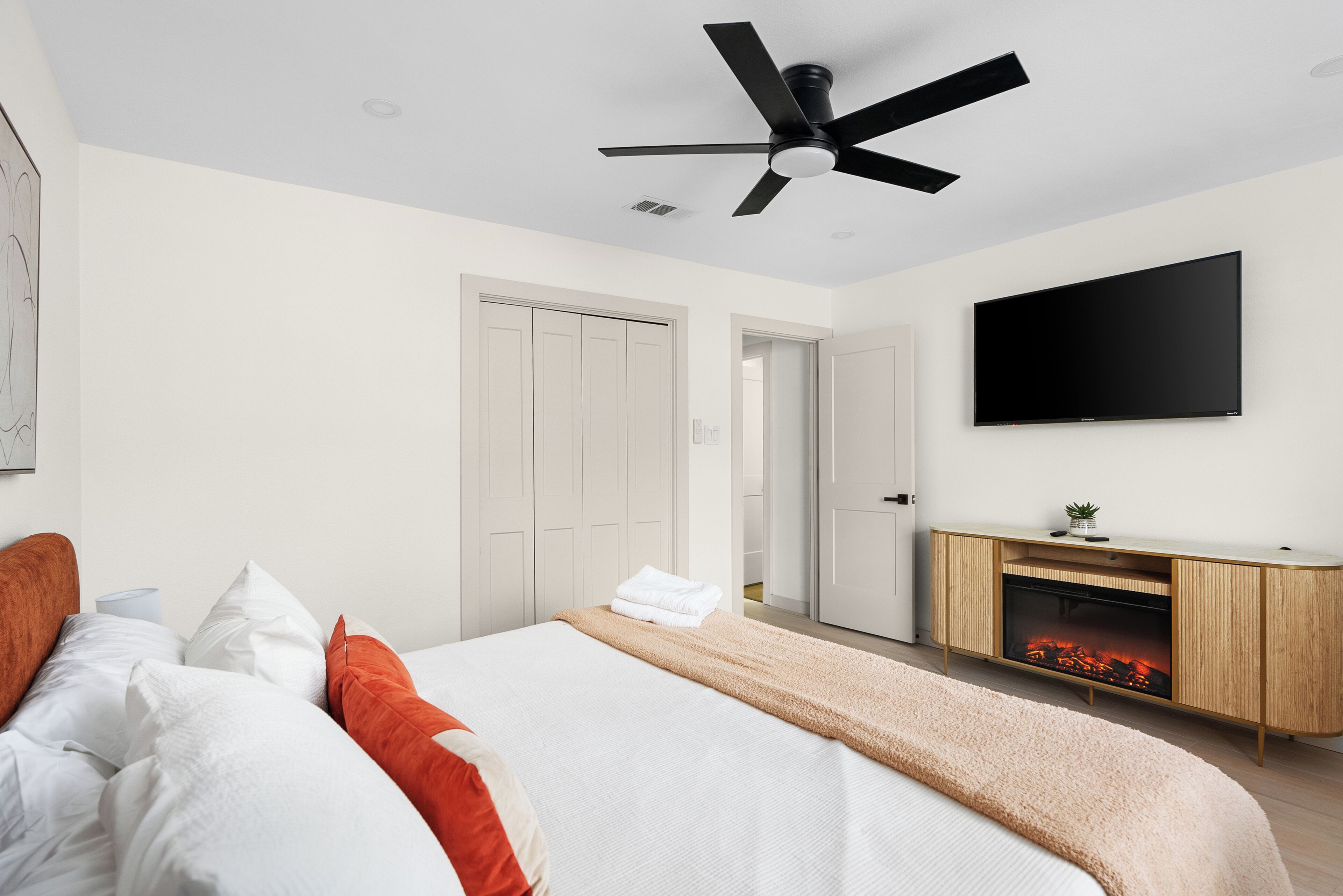 Une chambre à coucher moderne avec un lit, des murs blancs, un ventilateur de plafond, un téléviseur à écran plat et une cheminée.