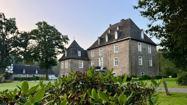 A large stone building with multiple windows and a prominent tower, surrounded by greenery.
