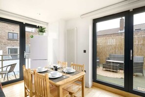 Un coin repas avec une table dressée pour quatre personnes, un réfrigérateur et une porte-fenêtre coulissante donnant sur un espace extérieur aménagé pour s’asseoir.