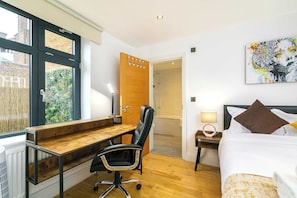 Ein Hotelzimmer mit einem Holztisch, einem schwarzen Bürostuhl, einem Bett mit weißen und braunen Kissen, einem Bild an der Wand und Blick auf ein Backsteingebäude durch ein Fenster.