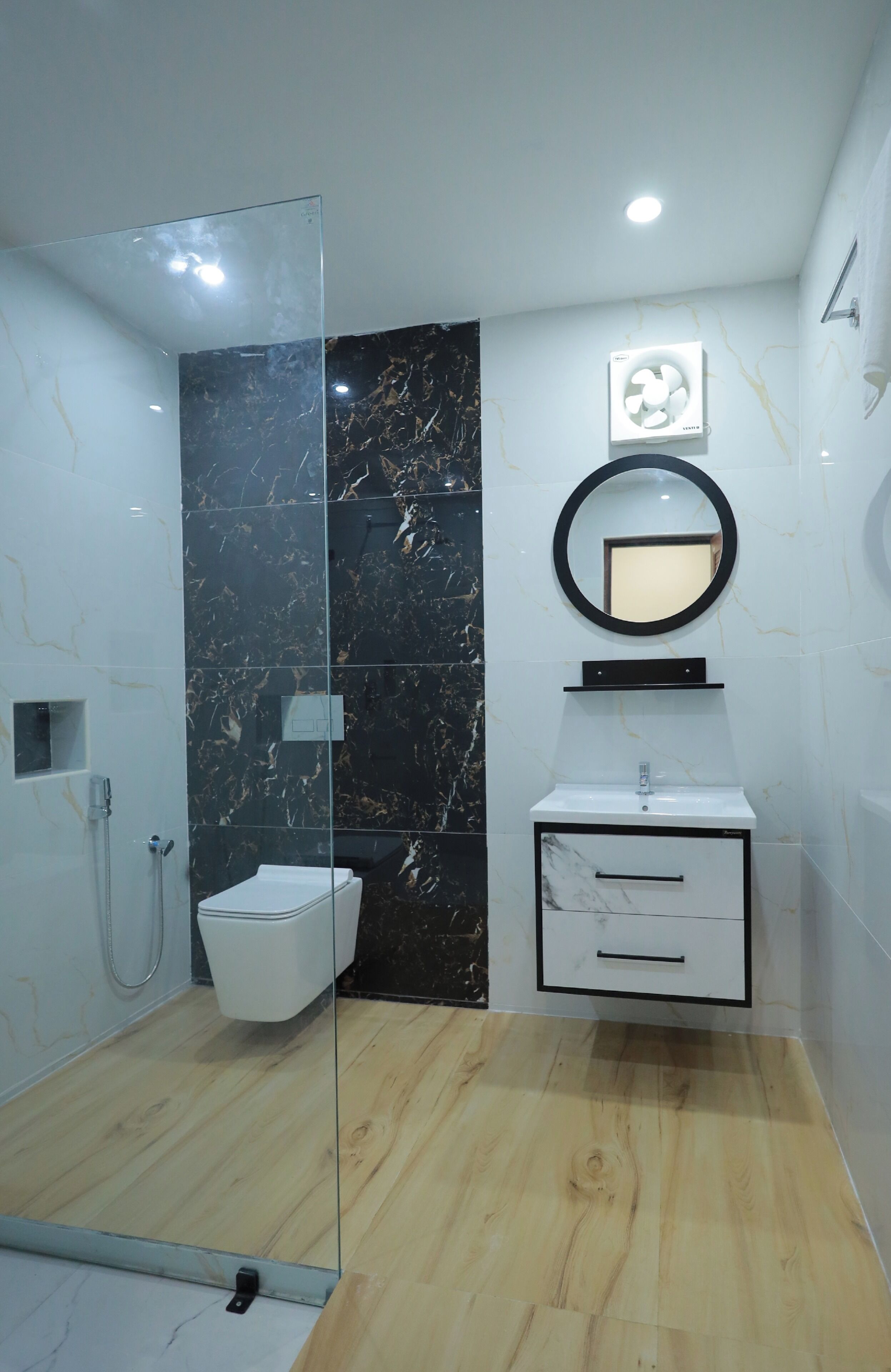 A modern bathroom with a glass shower enclosure, a marble wall, a white sink with a mirror, and a wooden floor.