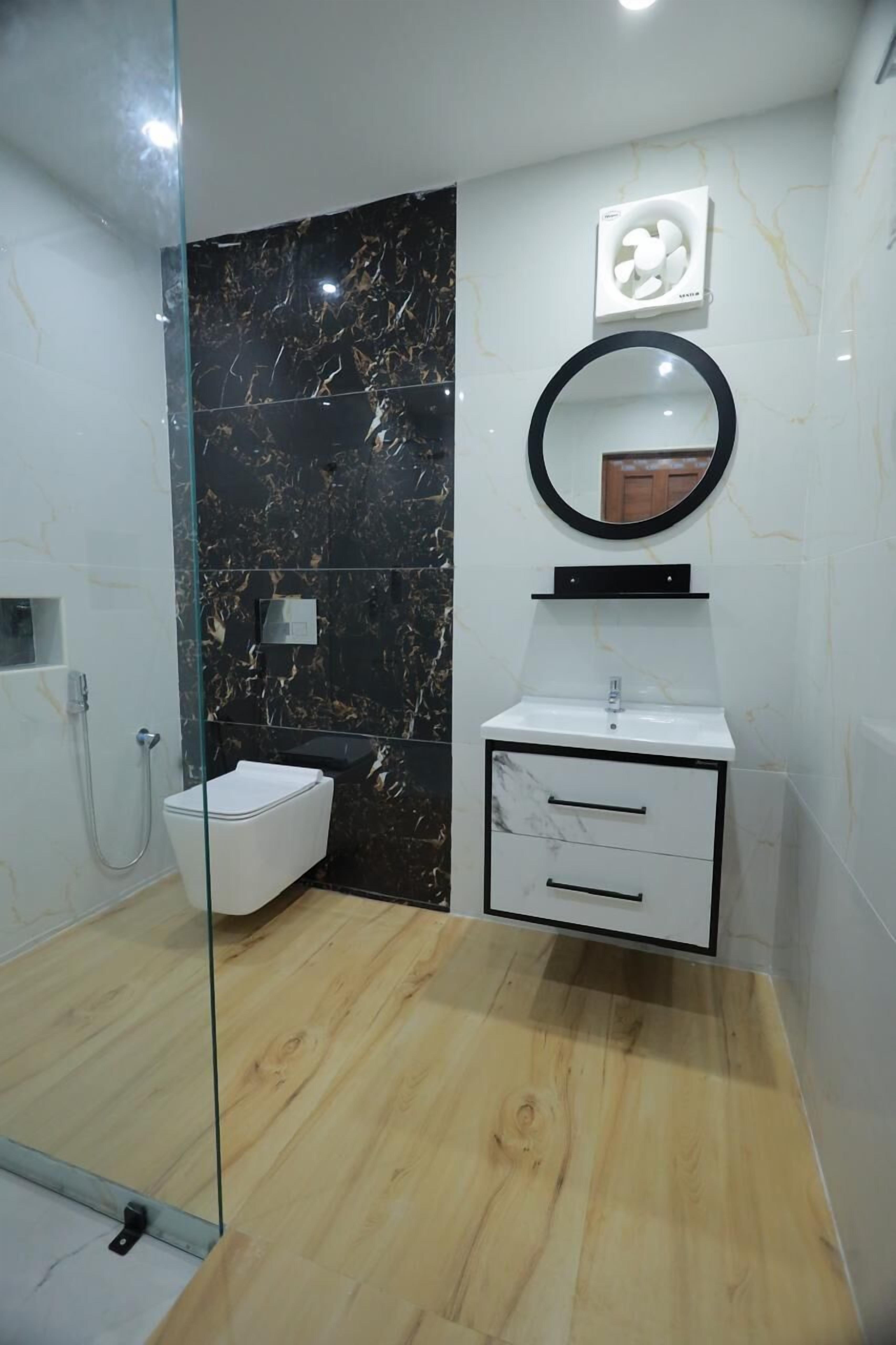 A bathroom with a glass shower enclosure, a white toilet, a marble backsplash, a white sink with a black cabinet, and a round mirror.