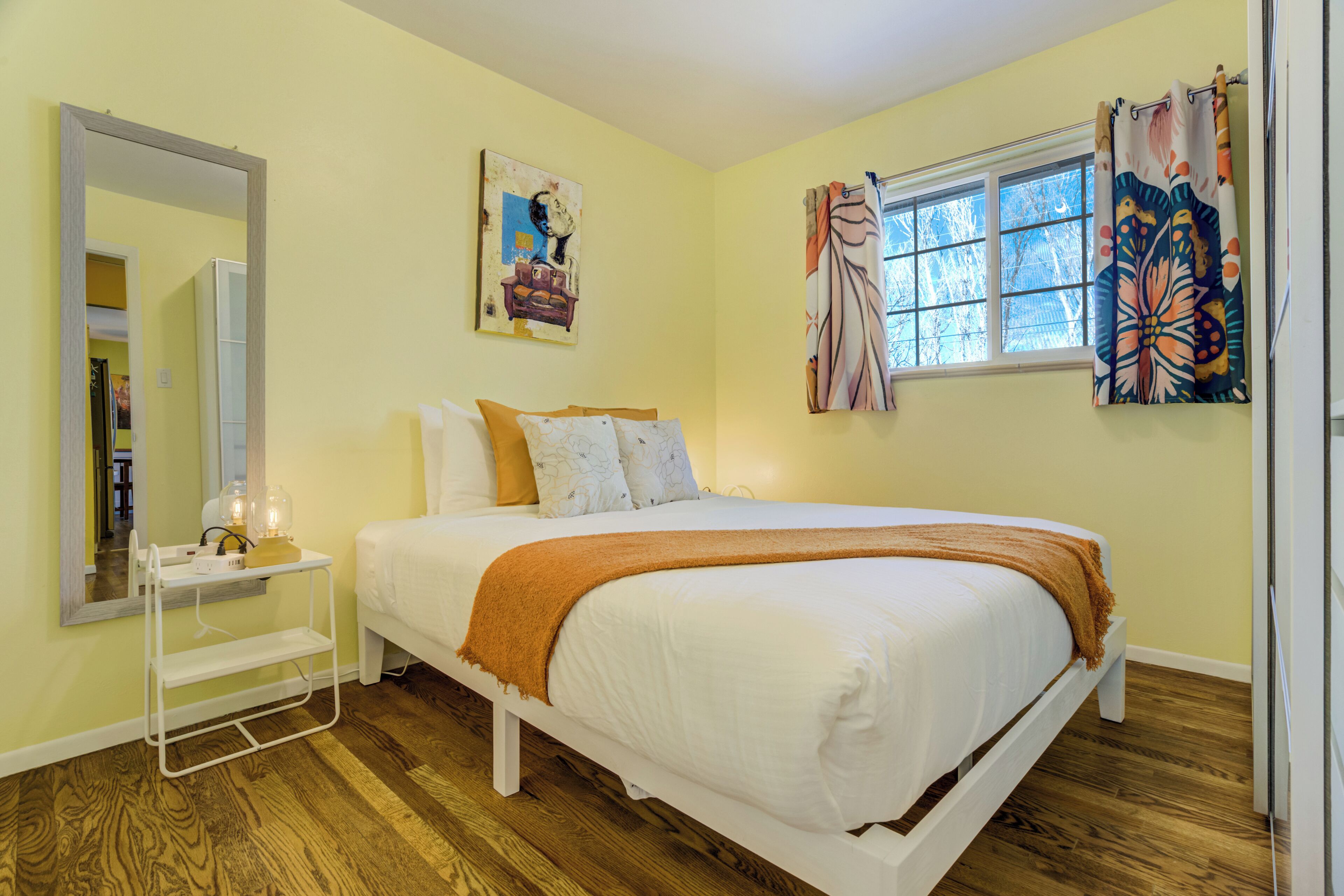 A bedroom with a bed, a bedside table, a mirror, and a window with curtains.