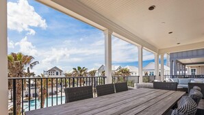 A covered outdoor area with a wooden table and wicker chairs, overlooking a swimming pool and palm trees. - Welcome to Tide and True | New Luxury Beachfront Retreat | Cottages at Romar 17 (Orange Beach)