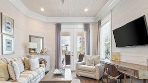 A living room with a sofa, armchair, coffee table, and a desk with a chair. There is a TV mounted on the wall and framed pictures on the wall. - Welcome to Tide and True | New Luxury Beachfront Retreat | Cottages at Romar 17 (Orange Beach)