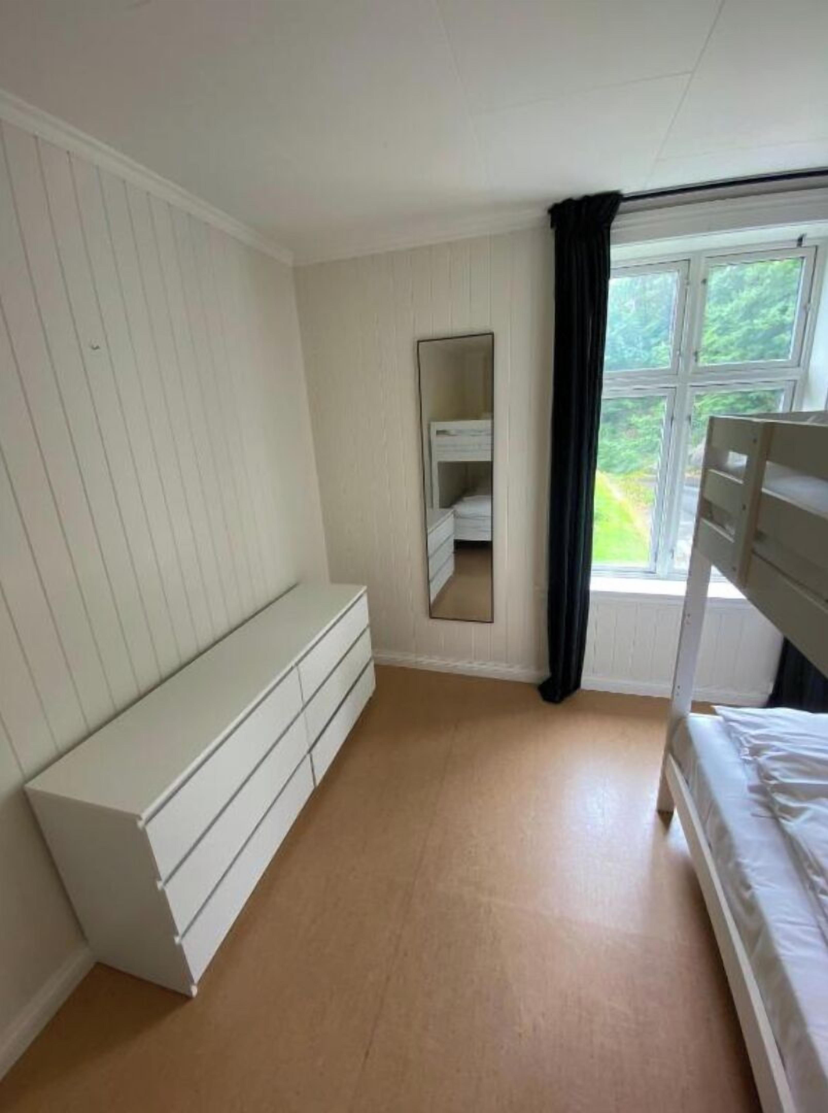 A bedroom with a bunk bed, a white dresser, a mirror, and a window with a view of greenery.
