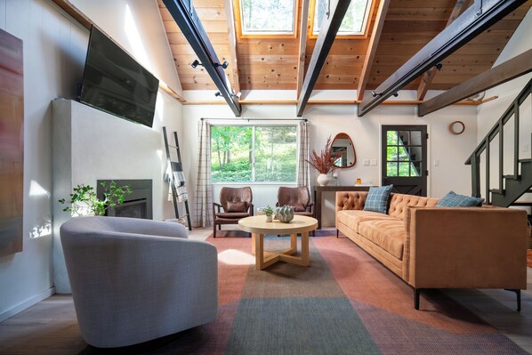 A modern living room with a fireplace, leather chairs, a sofa, and a large window offering a view of the outdoors. - The Robin: BLS Lake Access, Hot Tub, Curated Luxury Design, & Kids Game Room (Arnold)