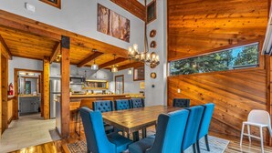 Un comedor amplio con una mesa de madera y sillas tapizadas en azul, una cocina al fondo y un ventanal con vistas a árboles.