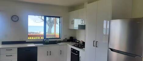 A modern kitchen with white cabinets, a stainless steel refrigerator, a dishwasher, and a window with a view of a tree and a fence.