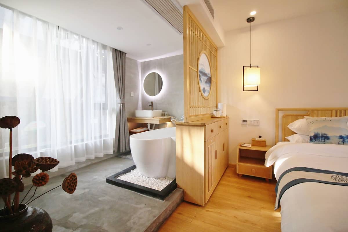 A hotel room with a bed, bedside table, nightstand, wooden dresser, round mirror, and a window with sheer curtains.