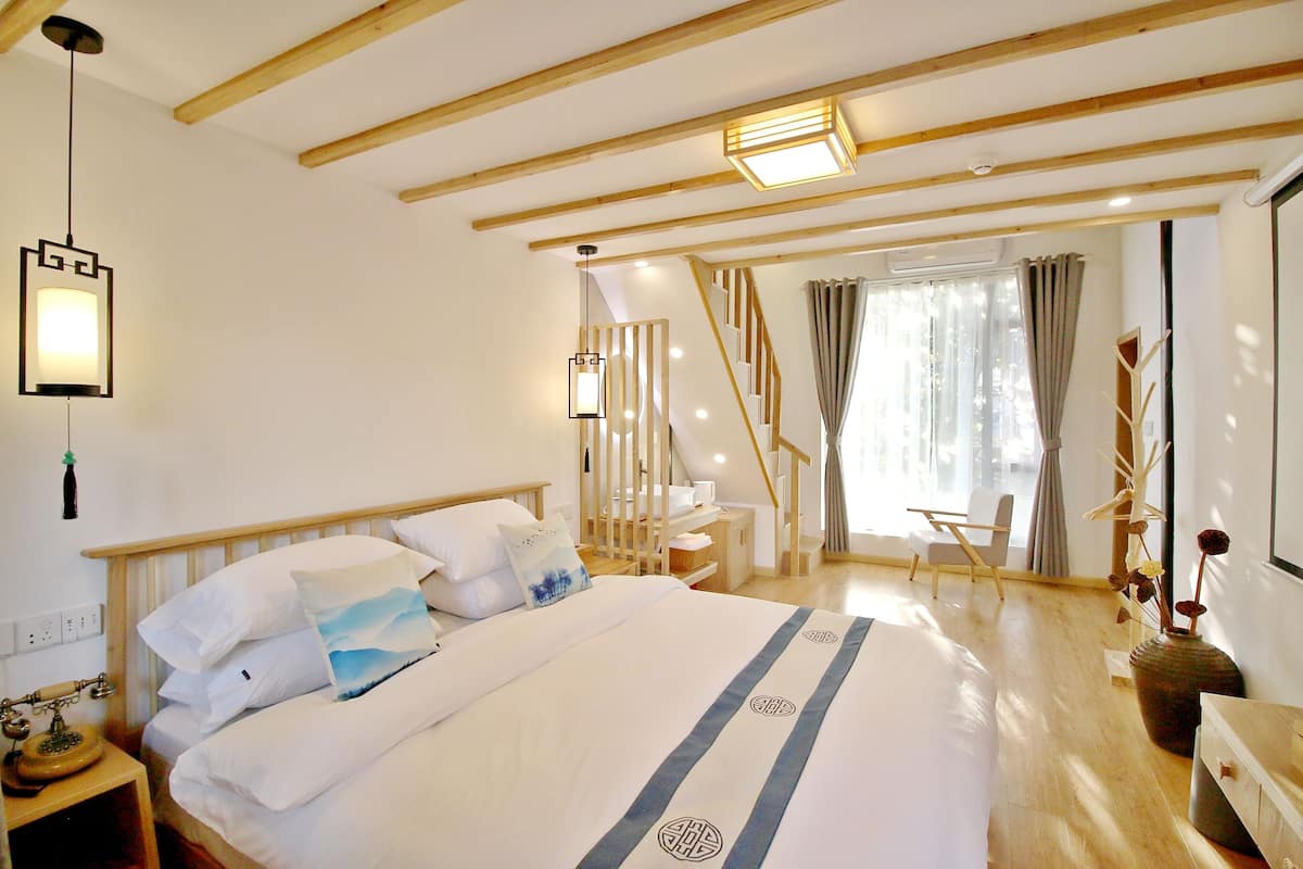 A bedroom with a large bed, wooden beams, a desk, a chair, and a window with curtains.