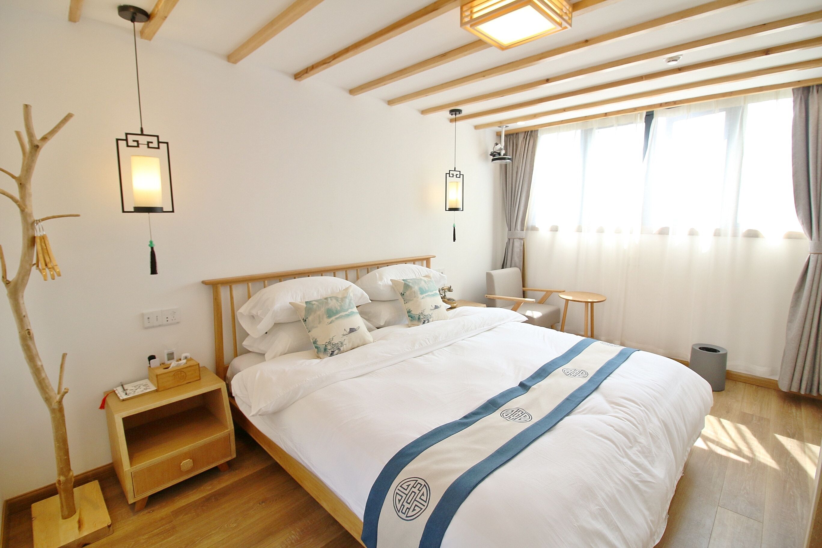 Quarto de hotel com cama de madeira, mesinhas de cabeceira, uma pequena escrivaninha e janela com cortinas.
