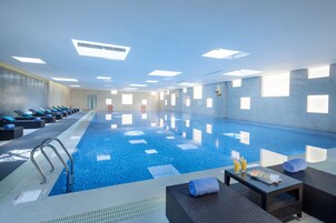An indoor swimming pool with clear water, sunlit ceiling, and lounge chairs along the side.