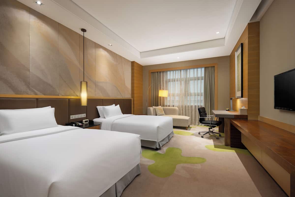 Ein Hotelzimmer mit zwei Betten, einem Schreibtisch, einem Stuhl, einem Fernseher und einem Fenster mit Vorhängen.