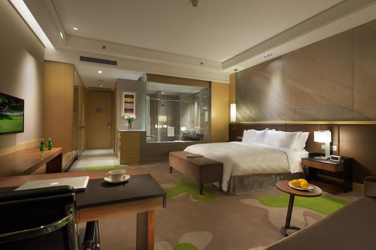 Ein Hotelzimmer mit Bett, Nachttisch, Lampe und einem kleinen runden Tisch mit Obst. Es gibt einen Schreibtisch mit Stuhl und einem Laptop, sowie ein Badezimmer mit Dusche und Spiegel.