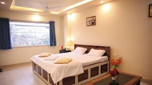 A bedroom with a large bed, a bedside table, a nightstand, a window with curtains, a ceiling fan, and a wall-mounted picture. - Hotel Kalpana (Patna)
