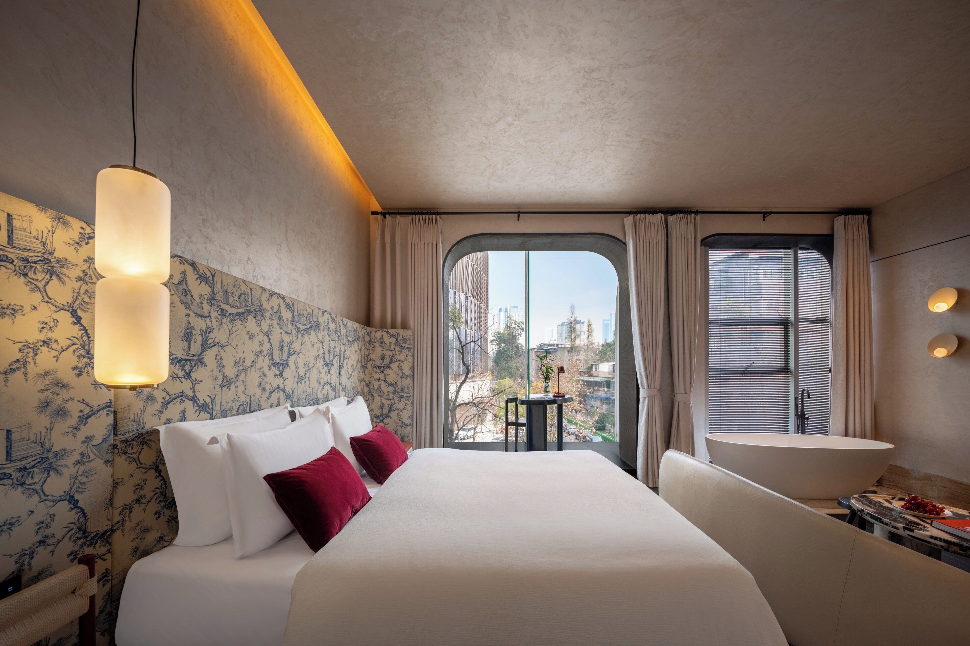 A hotel room with a large bed, a small table, a sofa, a large window with curtains, and a city view.