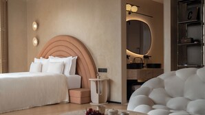 Una habitación de hotel moderna con una cama grande, una mesa de centro redonda con frutas, un sillón blanco y un diseño de cabecera único.