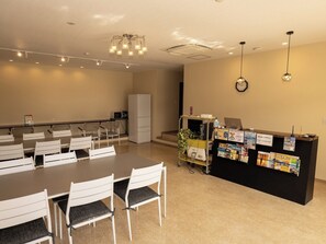 Eine Hotellobby mit einer Rezeption, einem Essbereich mit weißen Stühlen und einem braunen Tisch, einem Kühlschrank, einer Mikrowelle und einer Wanduhr.