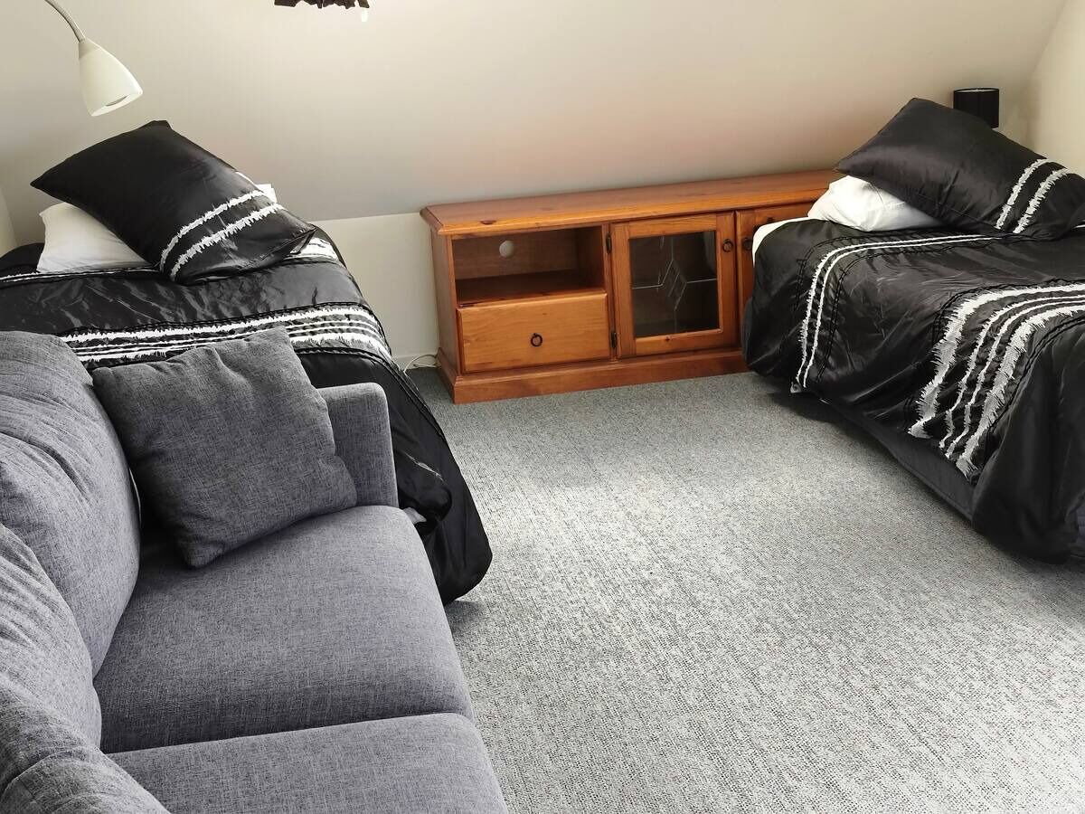 Una habitación con un sofá gris, dos camas negras con detalles blancos, un aparador de madera y una lámpara blanca.