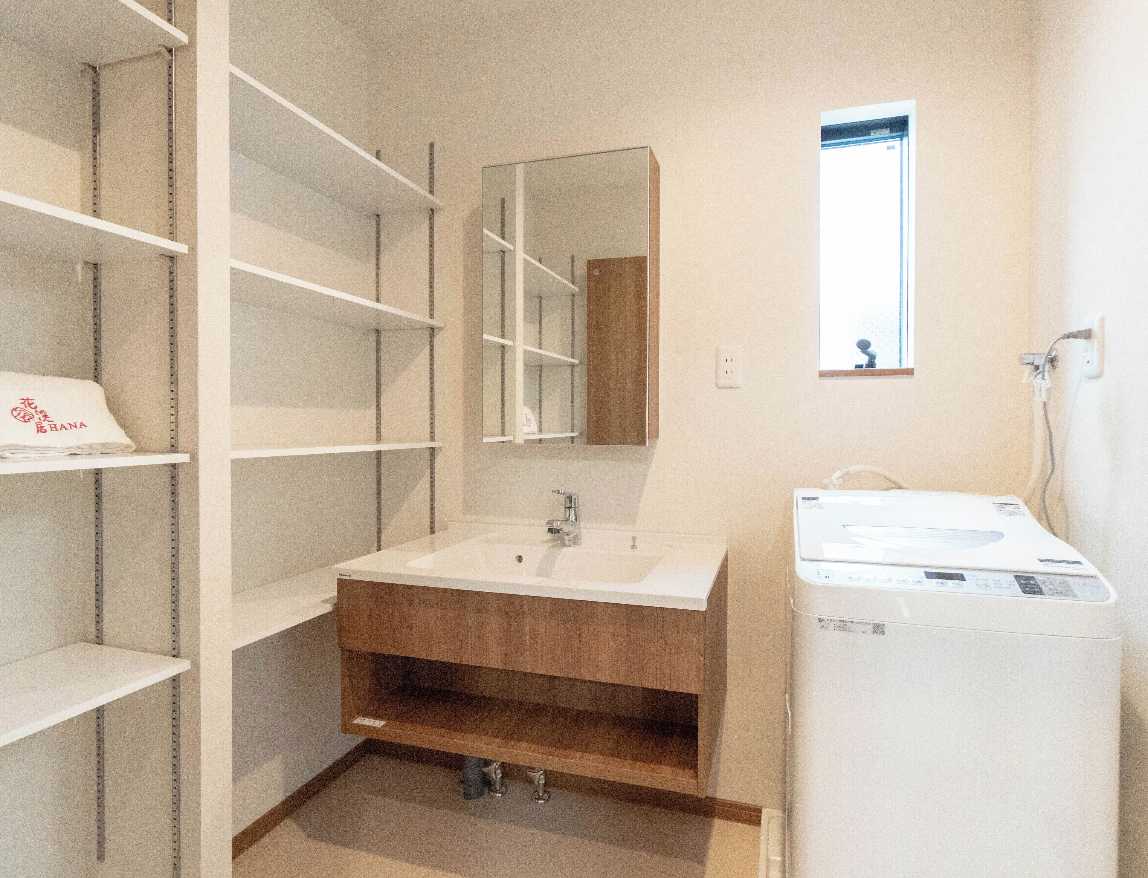 A bathroom with a white sink, mirror, and a white washing machine. There are shelves on the left and a wooden cabinet below the sink.