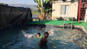 Une piscine où nagent deux personnes, entourée d’une zone abritée avec des chaises et une antenne parabolique.