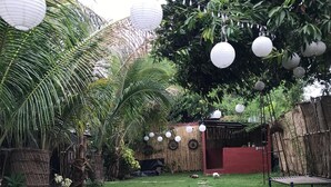 Un jardin avec un chemin de pierres menant à un bâtiment rouge, entouré d’une végétation luxuriante et illuminé de lanternes blanches en papier.