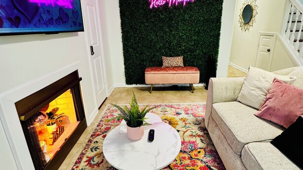 Ein modernes Wohnzimmer mit Kamin, rundem Couchtisch mit Pflanze, einer Couch mit Kissen, einem farbenfrohen Teppich und einem Neon-Leuchtschild an der Wand.