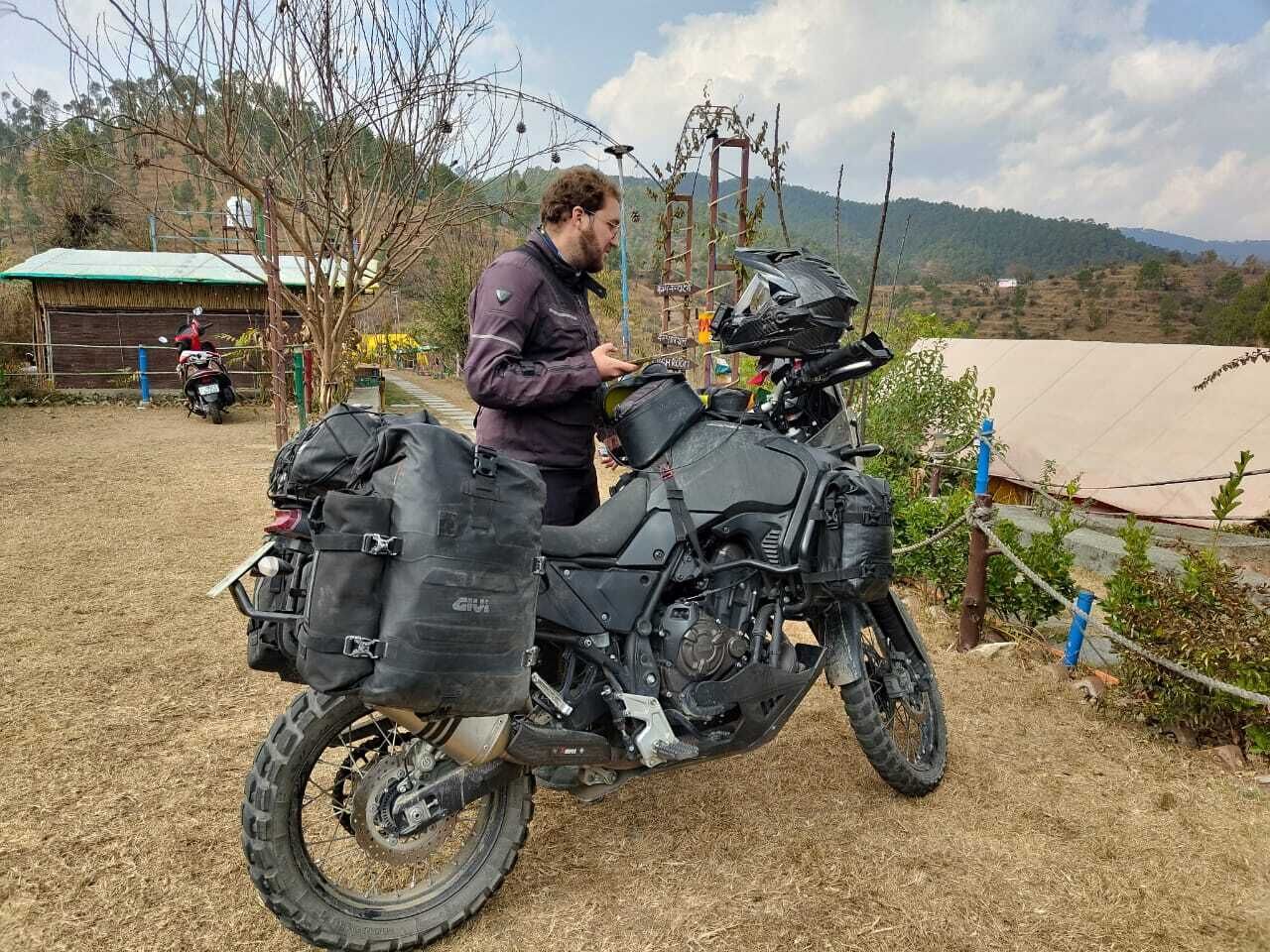 Una motocicleta con un depósito de carga grande, estacionada sobre terreno de tierra con un paisaje rural de fondo.