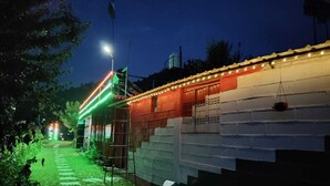 A pathway leading to a building with a brick facade and a string of lights along the roof edge. - Nisarg Camping Pods (Almora)