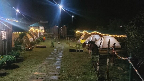 A rustic outdoor setting with tents and a small building, possibly a lodge or cabin, surrounded by a stone pathway and illuminated by string lights. - Nisarg Camping Pods (Almora)