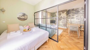 A bedroom with a bed, a glass door leading to a balcony, a dining area, and a wall with a nature-themed painting.
