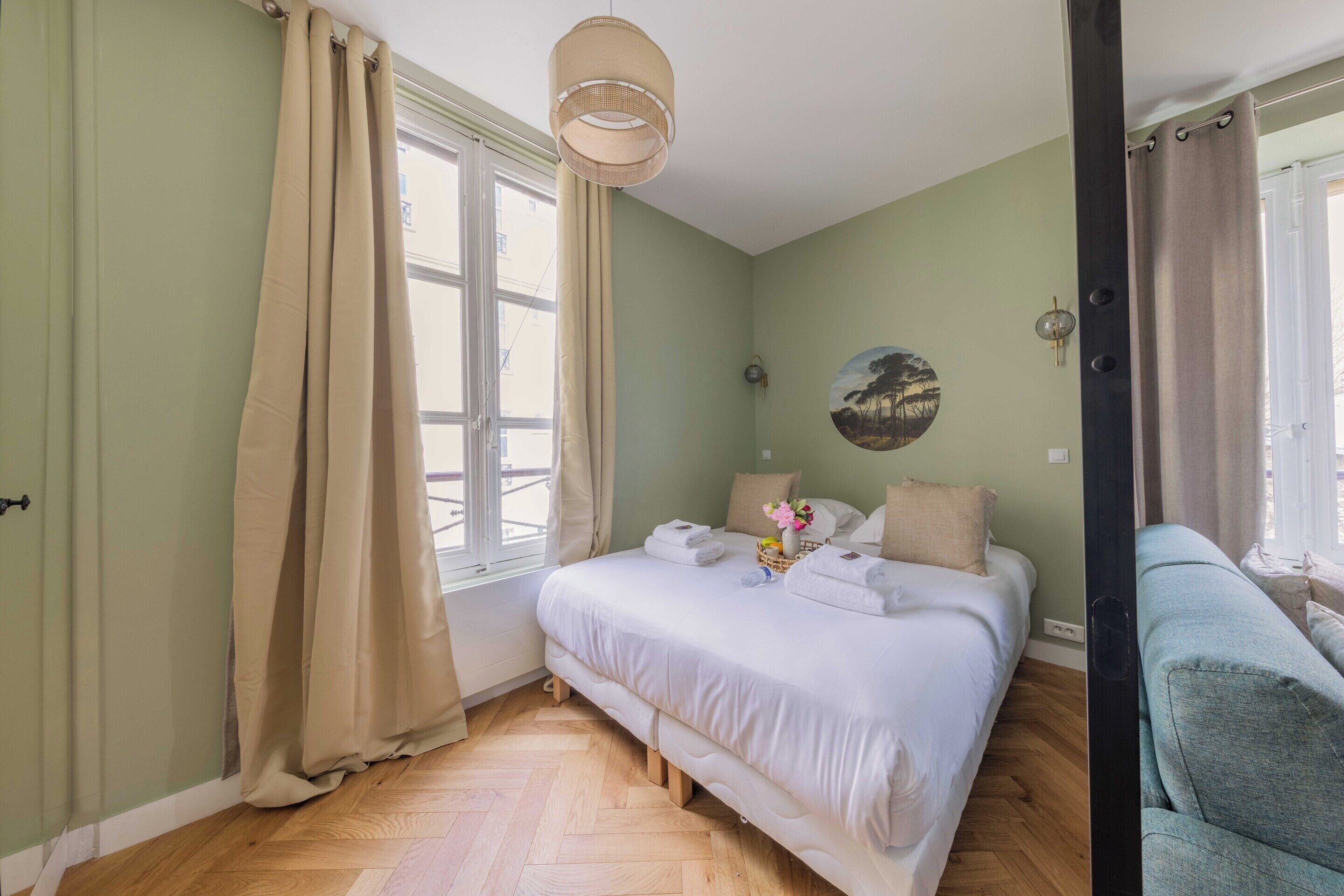 A bedroom with a bed, a sofa, a window with curtains, and a wall-mounted light fixture.
