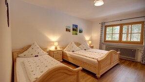 Ein Zimmer mit zwei Holzbetten, jeweils mit einem Nachttisch und einer Lampe. An der Wand hängen drei gerahmte Bilder, und ein Fenster mit Vorhängen.