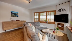 Ein modernes Wohnzimmer mit einer grauen Eckcouch, Holzboden, einer Holzkommode, einem kleinen runden Tisch mit einer Vase und einem an der Wand montierten Fernseher.