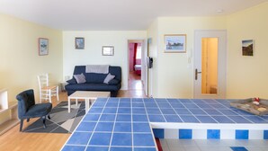 Una sala de estar moderna con una mesa de centro de azulejos azules, un sofá azul oscuro y una silla blanca. El suelo es de madera y hay una puerta que da a otra habitación.