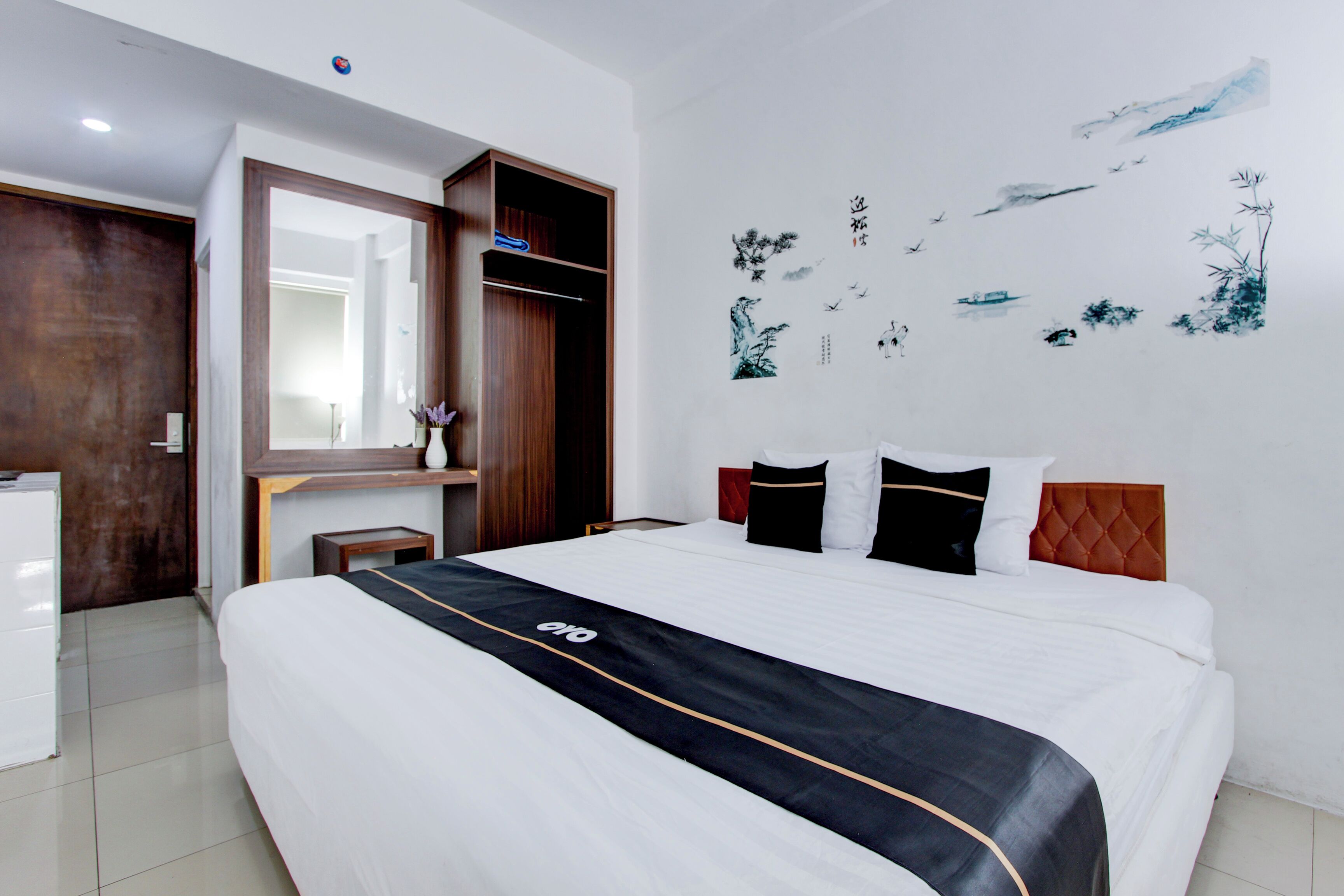 A hotel room with a large bed, a wooden wardrobe, a small table with a vase, and a wall with artwork.