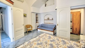 A bedroom with a bed, a sofa, a chair, and a small table. There are two doors, one leading to a hallway and the other to a bathroom. - Appartamento Attilia B: A graceful apartment which faces the sun and the sea, with Free WI-FI. (Amalfi)