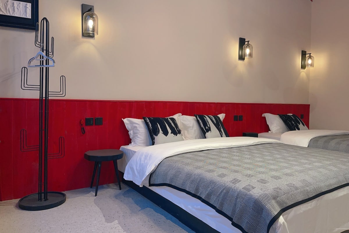 A hotel room with a red accent wall, two beds with patterned bedding, a standing lamp, and a coat rack.