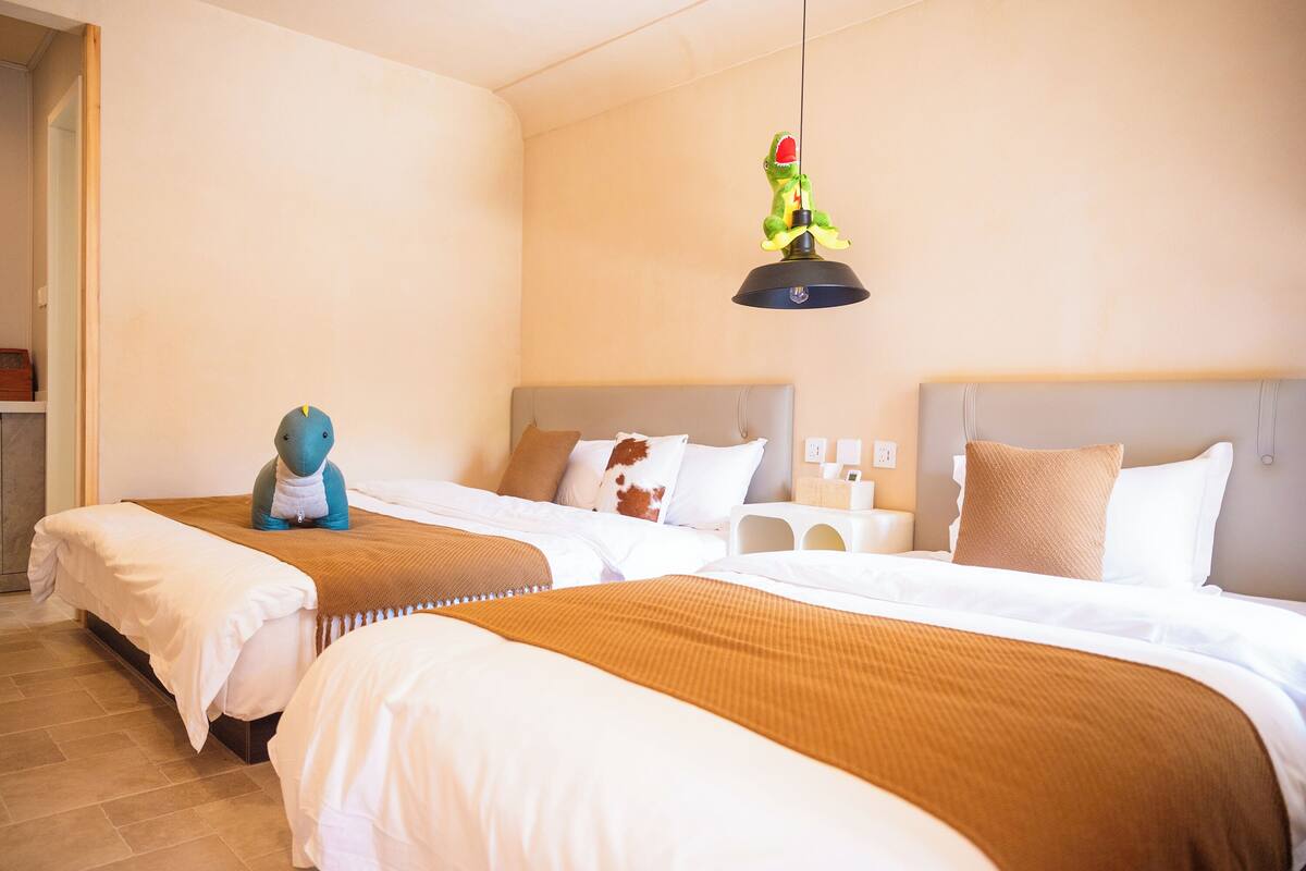 A hotel room with two beds, a hanging light fixture, and a small table with a lamp.