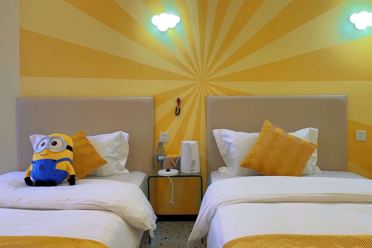 A hotel room with two beds, each with white bedding and a yellow pillow. There is a Minion plush toy on one bed. The room has a yellow and orange wall design with radial light fixtures.