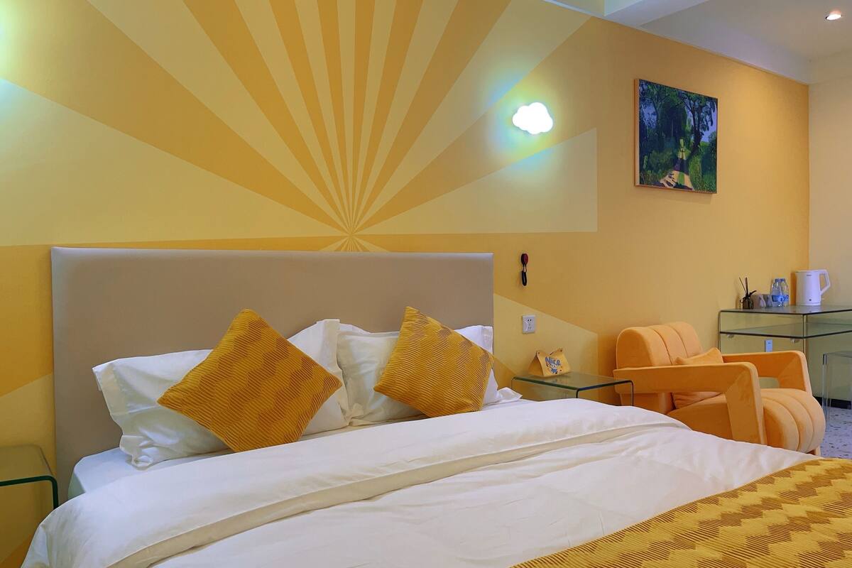 A hotel room with a bed, a desk, a chair, a painting on the wall, and a nightstand with a lamp.