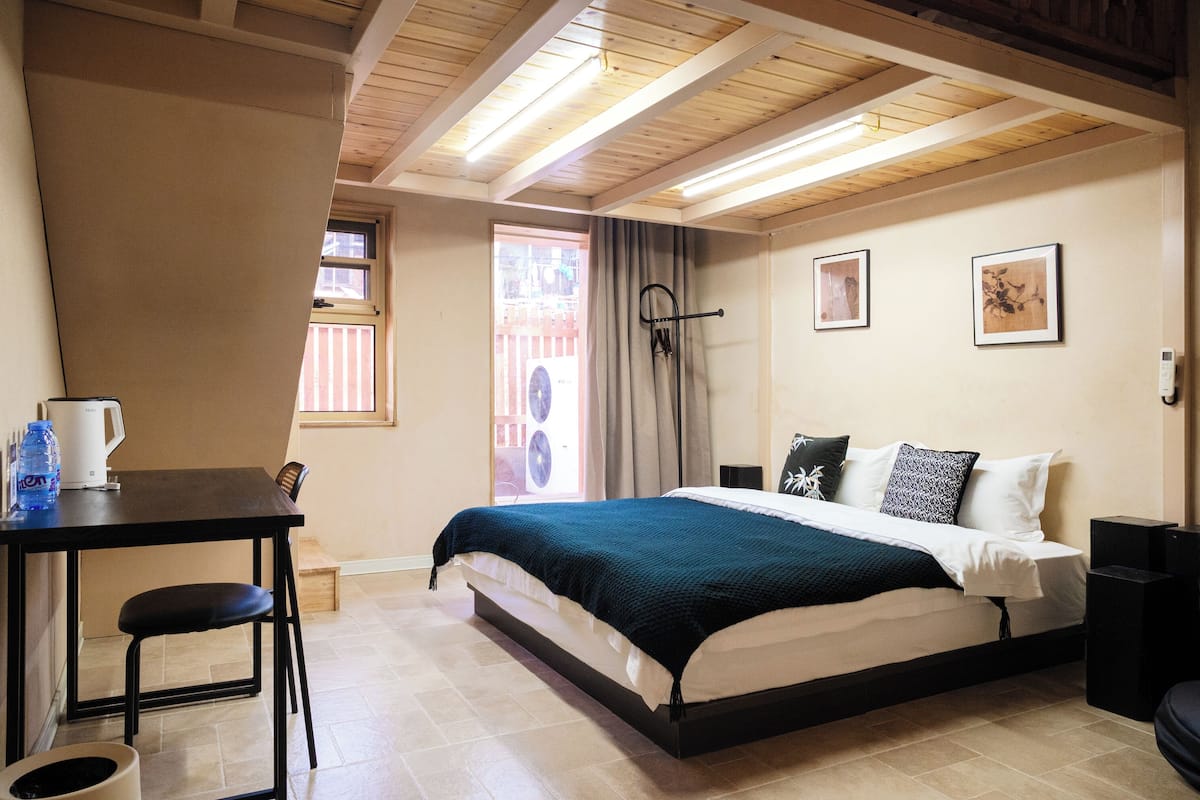 A bedroom with a bed, a table with a chair, a window with curtains, and a fan.