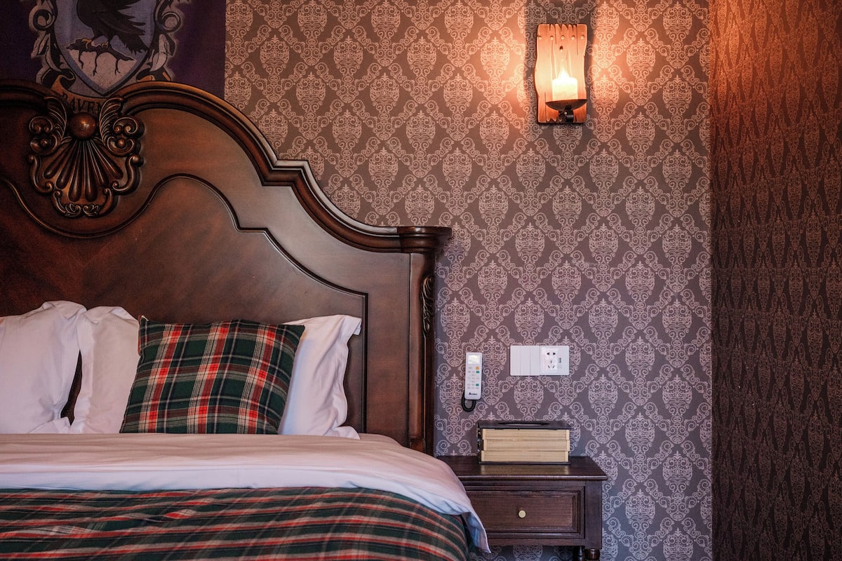 A neatly made bed with a plaid blanket, a wooden headboard, a bedside table with a book, and a wall-mounted telephone.