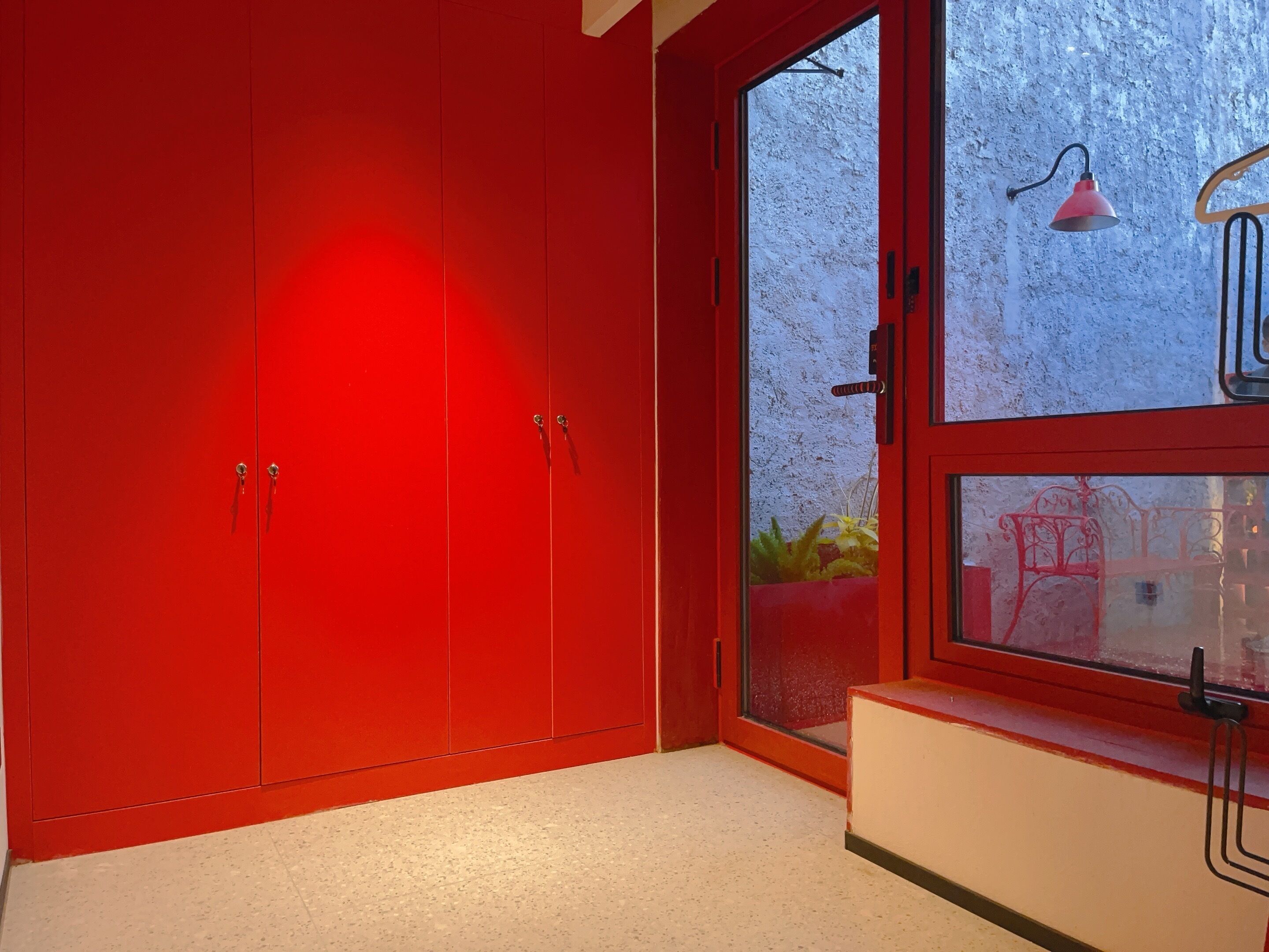 Una habitación con un armario rojo, una ventana con vistas a plantas y una lámpara roja.