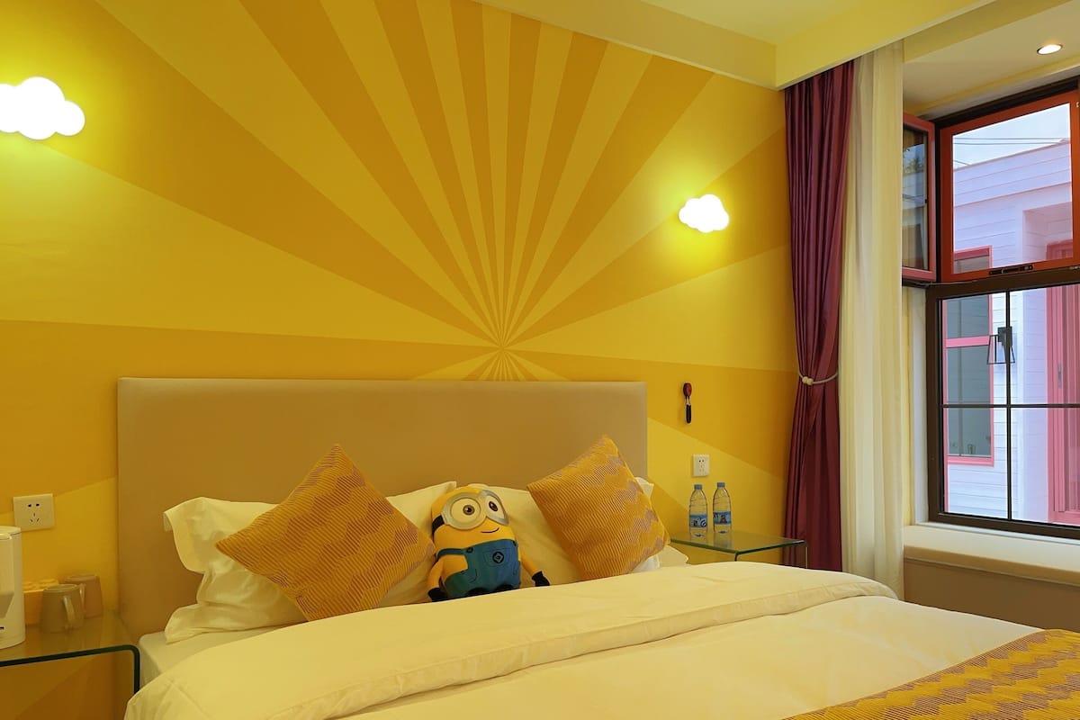 A hotel room with a bed, a Minion plush toy, a bedside table with water bottles, and a window with curtains.
