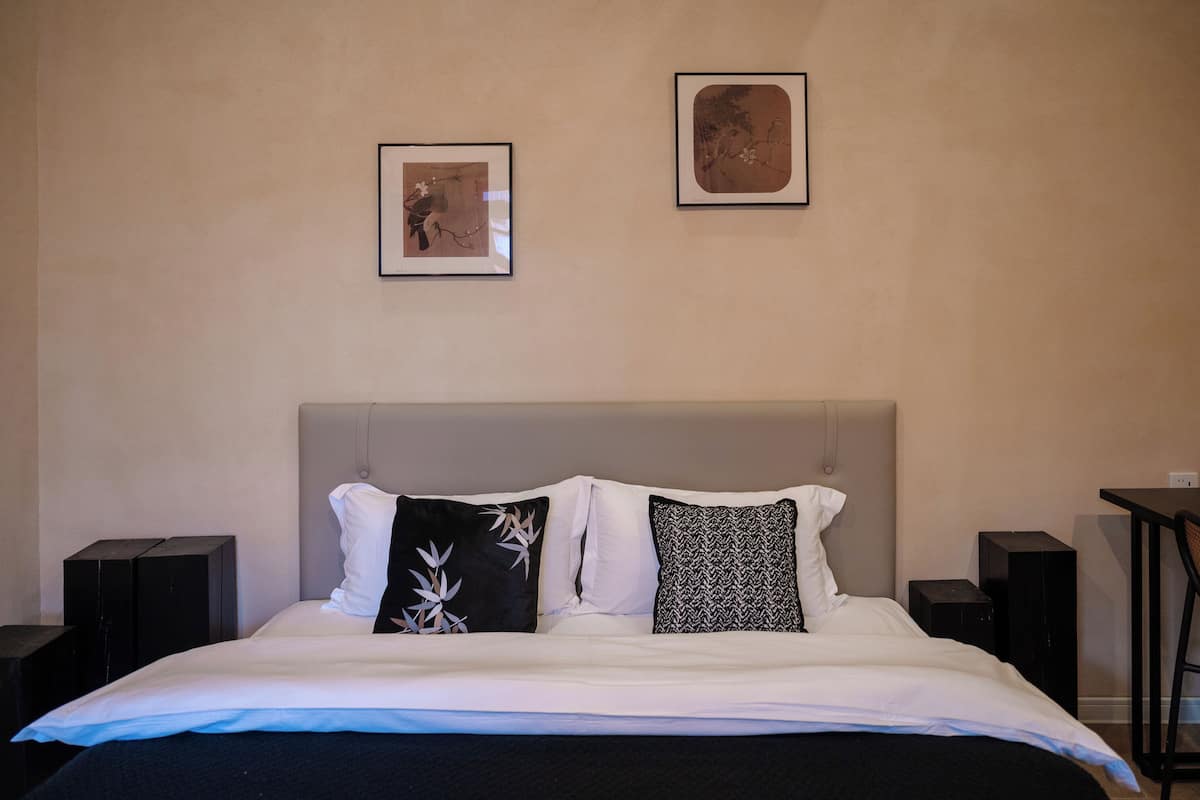 A hotel room with a bed, two framed pictures on the wall, a grey headboard, and two speakers on either side of the bed.